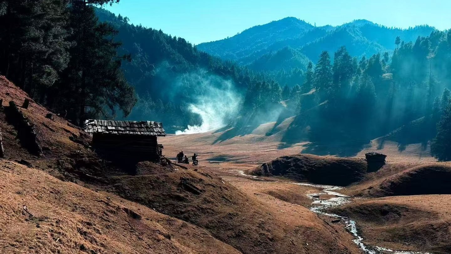 A wooden hut beside a mountain stream in a sunlit valley, with mist rising through pine forest.