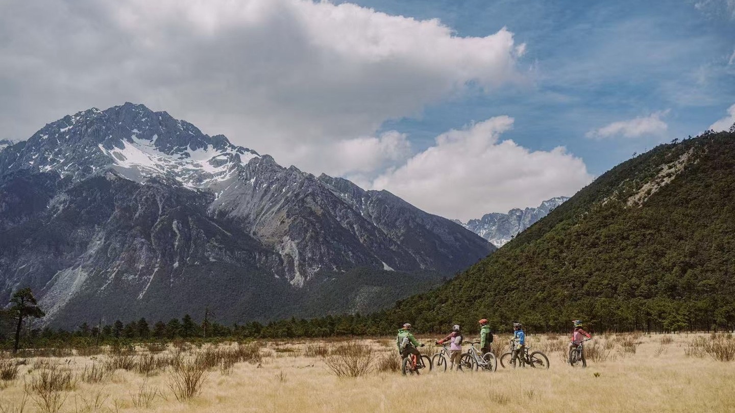 A group of mountain bikers pauses in a golden dry-grass meadow beneath the snow-streaked ridges of Jade Dragon Snow Mountain.
