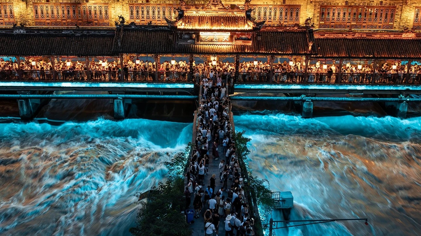 The illuminated Nanqiao Bridge and covered walkways at Dujiangyan above the turquoise Min River at dusk.