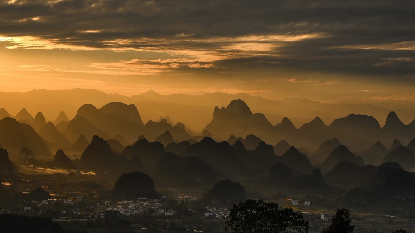 Rows of karst peaks fading into golden haze at sunset above a valley of fields and villages