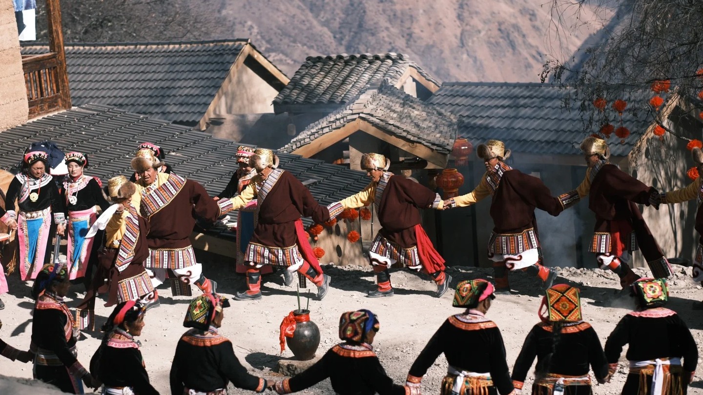 Villagers in traditional Jiarong Tibetan dress dancing in a circle in a stone-built village square.