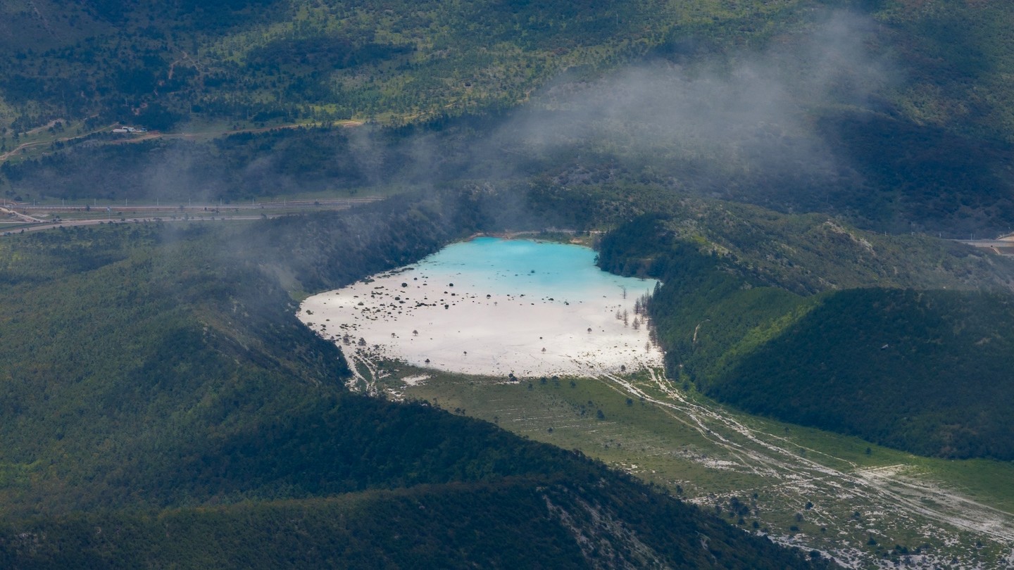 Aerial view of a turquoise alpine lake on a white sand basin, ringed by dark forested ridges and drifting mist.