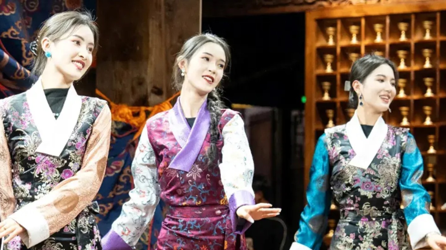 Three women in traditional Tibetan silk dresses perform a cultural dance on a stage.