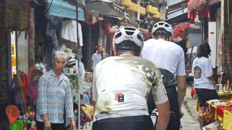Two cyclists in professional gear ride through a narrow, traditional Chinese market street with local vendors and lanterns.