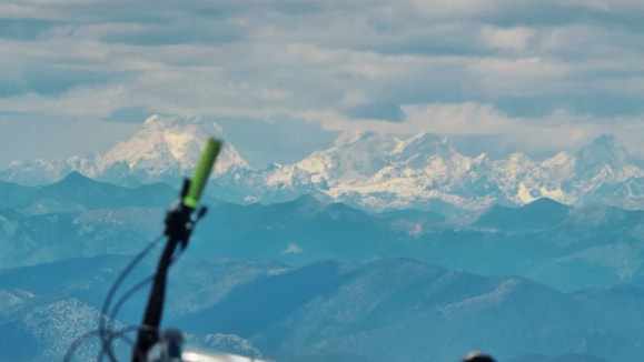 A mountain bike rests on a gravel summit with a distant snow-capped range under heavy clouds.