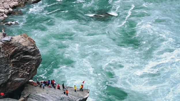 Tourists standing on a rocky viewing platform overlooking the rushing turquoise waters of Tiger Leaping Gorge.