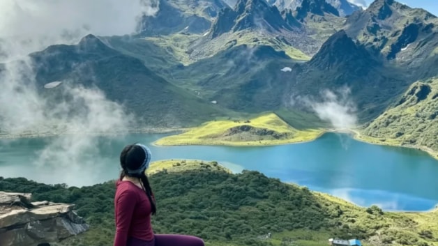 A female hiker sits on a cliff overlooking a blue alpine lake and majestic snowy mountain peaks.
