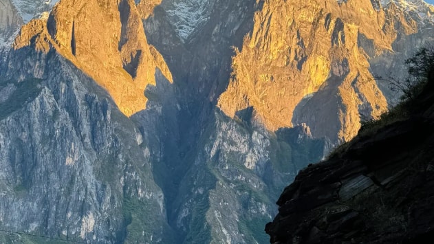 A lone hiker on a narrow gorge trail below jagged peaks lit gold by the rising sun.