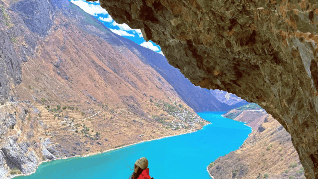 A hiker in a red jacket stands under a rock overhang high above the turquoise river winding through Tiger Leaping Gorge.