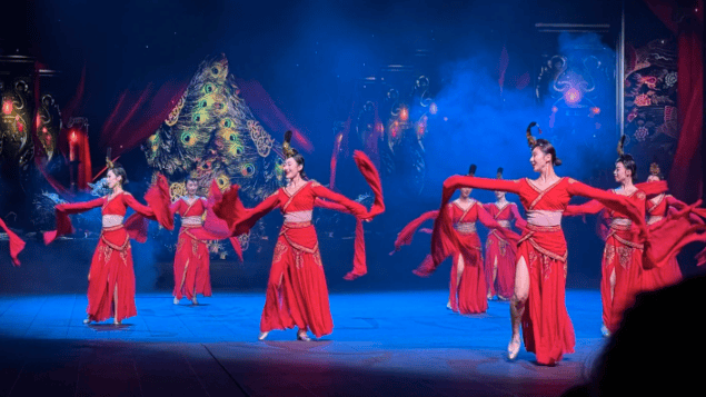 Dancers in vibrant red traditional Chinese costumes perform a folk dance on a stage with blue lighting.
