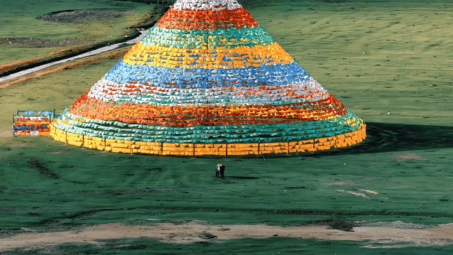 A conical structure draped in colorful Tibetan prayer flags stands on a green grassy plain near a river.
