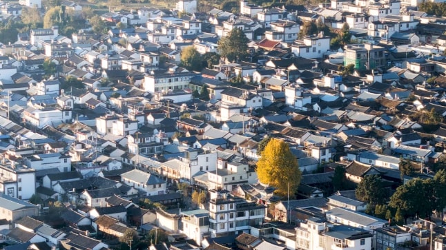 Aerial view of a dense village of white-walled, grey-tiled courtyard houses in early morning light, terraced fields on the slope above.