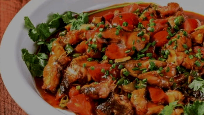 A white bowl of spicy Chinese braised fish with tomatoes, chives, and cilantro on a wooden table.
