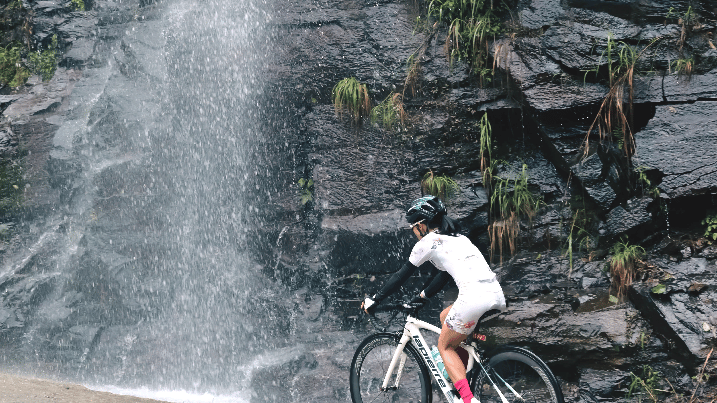 A female cyclist on a white road bike riding past a scenic waterfall on a rocky mountain road.