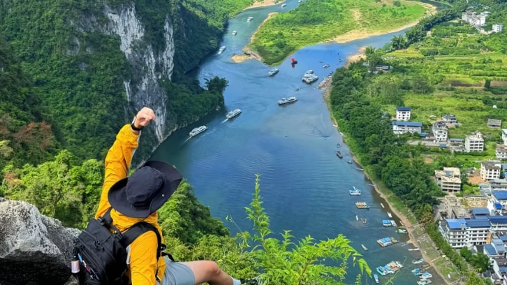 A hiker overlooks the Li River and limestone karst mountains in Guilin, China, from a rocky summit.