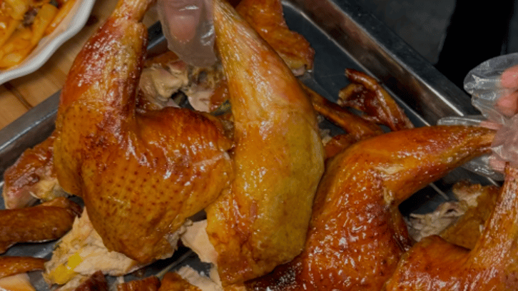 A person wearing plastic gloves holds up a crispy roasted whole chicken leg over a metal platter.