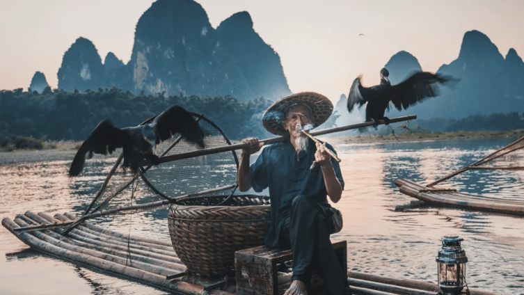 A traditional cormorant fisherman smoking a pipe on a bamboo raft at sunset in Yangshuo, China.