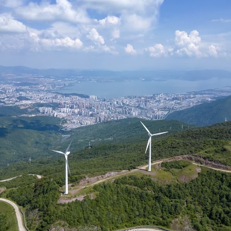 Aerial view of a gravel road winding past wind turbines on green ridges, Dali and Erhai Lake beyond
