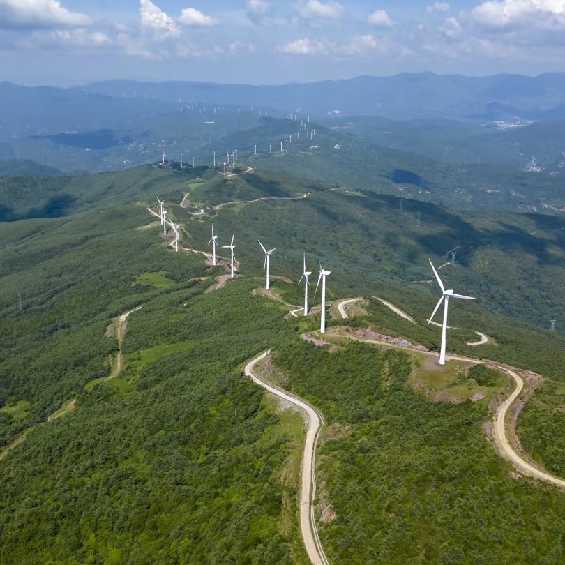 Aerial view of a dirt road snaking along a forested ridge lined with wind turbines