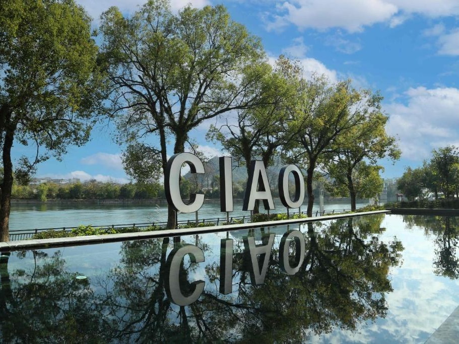 Steel CIAO letters on a mirror pool reflecting trees and sky, the Li River behind