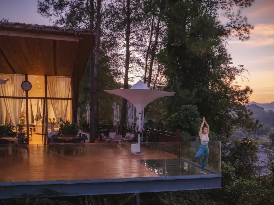 Timber pavilion beside a glass-edged deck jutting over a wooded slope at sunset, karst peaks in the distance