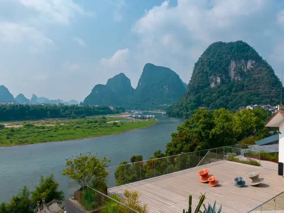 Timber rooftop deck with low chairs behind glass railing, Li River and karst peaks beyond
