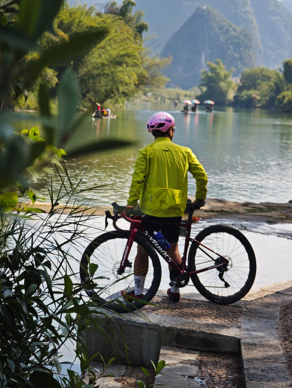 Rider in a cycling jacket standing with a bike on a stone landing, watching bamboo rafts drift down the Yulong River below karst peaks