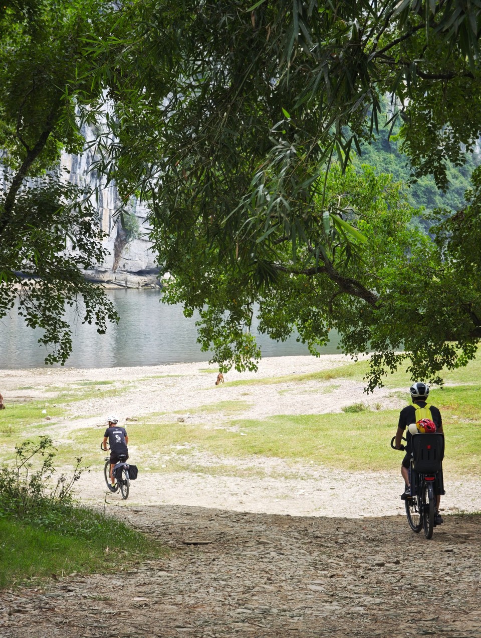 Two riders on a riverside track under trees near Yangshuo, one on an e-bike with a child seat fitted on the rear rack