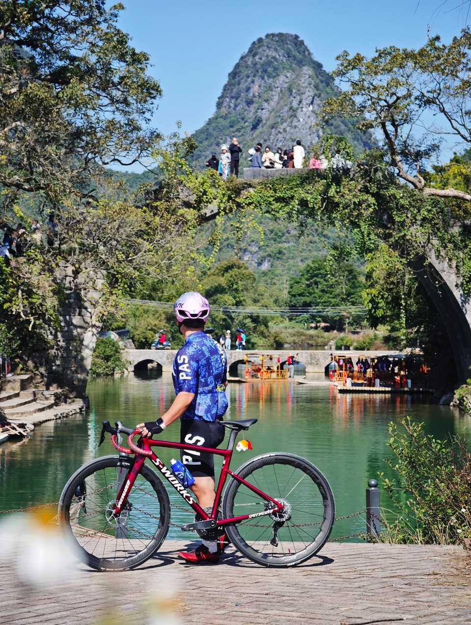 Rider with a road bike on the riverside path below an ivy-covered Ming-era stone arch bridge on the Yulong River, a karst peak behind
