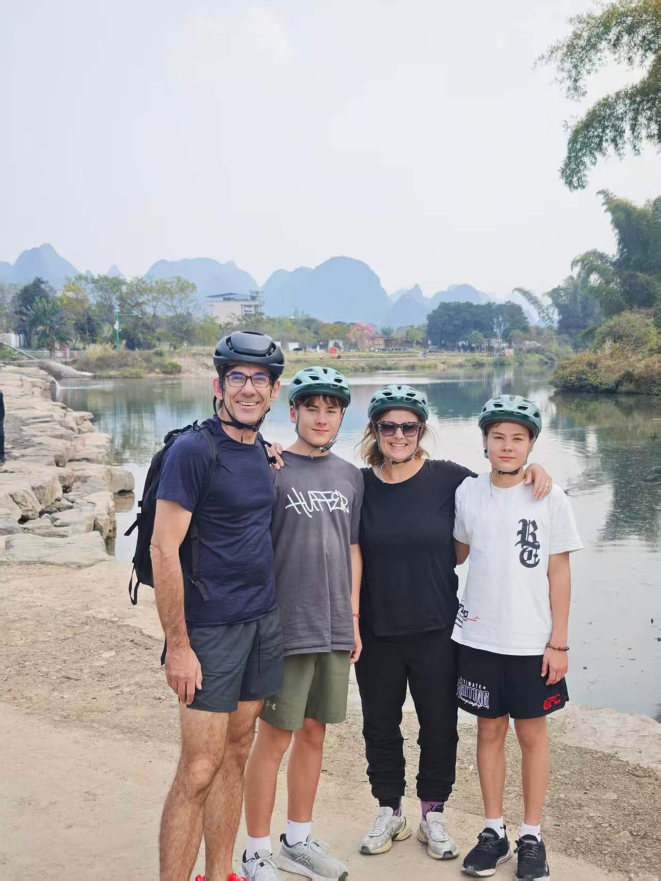 Family of four in cycling helmets on the Yulong riverbank with karst peaks in the haze behind
