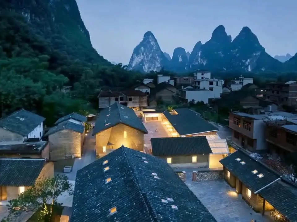 Grey-tiled village houses lit from within at dusk, karst peaks rising behind