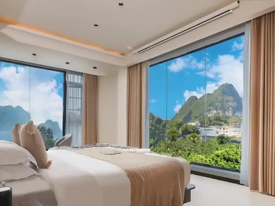 Bed between two floor-to-ceiling windows looking onto green karst peaks and a village